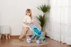 A young girl is pushing a blue and teal toy stroller with a Stitch plush toy inside, in a room with a white brick wall and a potted plant. Náhľad obrázku produktu