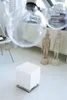 A white cube-shaped humidifier sits on a white floor in a bright, minimalist room. Two decorative statues stand behind it, and a chair is visible to the left. Produktbild-Vorschau