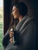 A woman wrapped in a blanket holds a travel mug while looking out a window. Anteprima dell’immagine prodotto
