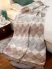 A blanket draped over a bed, with a bedside table and lamp visible. Produktbild-Vorschau