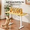 A cat is interacting with a standing desk, with large gold letters spelling '70KG' on the desk surface. Produktbild-Vorschau