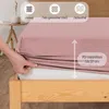A hand is shown stretching a pink fitted sheet over a mattress on a wooden bed frame. Icons and text describing the product's features are overlaid at the top and side. Produktbild-Vorschau