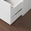 A close-up view of a white drawer with a metal slide mechanism, resting on a brown wooden floor. Produktbild-Vorschau