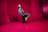 A woman sits on a black rolling stool in a bright pink room. Produktbild-Vorschau