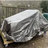 A silver tarp covering an object on a wooden pallet with wheels, in an outdoor setting with a wooden fence and trash cans in the background. Produktbild-Vorschau