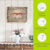 A wall clock with a scenic image is displayed on a white paneled wall, with flowers in a vase on a dresser below. To the right, there are marketing icons and text. Produktbild-Vorschau