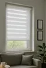 A window with a white zebra blind is shown, with a grey sofa and a plant in the foreground. Produktbild-Vorschau