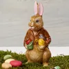 A ceramic Easter bunny figurine dressed in a brown jacket, holding a yellow Easter egg and a green bucket with a red lid, standing on moss with colorful eggs around it. Náhled obrázku produktu