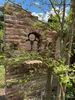 A weathered brick wall with a decorative iron window frame, partially obscured by green foliage and tree branches. Produktbild-Vorschau