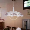 A pendant lamp with a floral pattern hangs over a dining table in a room with wooden furniture and a window. Náhled obrázku produktu