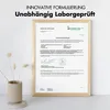 A framed certificate of analysis is displayed on a light-colored surface, with decorative dried flowers in a ribbed vase to the left. Produktbild-Vorschau