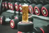A beer mug with a muscular arm handle sits on a dumbbell in a gym setting, with rows of dumbbells in the background. Produktbild-Vorschau