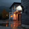 A solar LED floodlight with a motion sensor is mounted on the exterior wall of a house at night, illuminating a wet driveway. Produktbild-Vorschau
