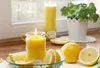 A lit candle and lemons are arranged on a wooden surface near a window, with a potted plant in the background. Produktbild-Vorschau