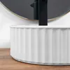 A close-up shot of a white ceramic washbasin with vertical fluting, resting on a wooden surface. A black faucet and a round mirror are visible in the background. Produktbild-Vorschau