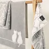 Three towels are displayed on a wooden rack. The largest towel in the center has a pattern of two white cats sitting on a line. The towel to the left is folded and has a similar cat pattern. A smaller towel hangs on the right, also with a cat pattern and a Produktbild-Vorschau