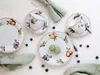A top-down view of a fruit-themed porcelain tea set with two cups and saucers, three plates, and two macarons, arranged on a white background with scattered blueberries and green napkins. Náhľad obrázku produktu