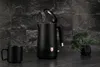 A black electric kettle with its lid open, a black mug, and a small potted plant are displayed on a dark countertop against a textured dark gray wall. Náhľad obrázku produktu