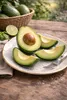Avocado slices and a whole avocado on a plate, with a basket of avocados and a lime in the background, set on a wooden table. Náhled obrázku produktu