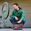 A man is using a portable air compressor to inflate a bicycle tire. The bicycle is mounted on a stand. Voir les images du produit