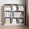 A white, multi-compartment bookshelf filled with books, boxes, and decorative items, set against a light gray wall and a beige curtain. Produktbild-Vorschau