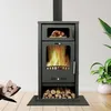 A black wood-burning stove with a glass door showing flames, a top compartment with a roasted chicken, and a bottom shelf with firewood. The stove is placed on a wooden floor in front of a white wall, with a potted plant to the right and a pile of firewood Produktbild-Vorschau