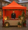 A woman stands behind a table at an outdoor market stall decorated for Christmas, selling jars of preserves. Podgląd zdjęcia produktu