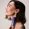 A woman is holding a bottle of serum, looking to the side with a smile. Produktbild-Vorschau