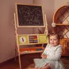 A young child is sitting on the floor in front of a wooden easel with a chalkboard and educational toys. Produktbild-Vorschau