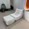 A white sofa with a matching ottoman is placed in a modern living room with a fireplace and a floor lamp. Produktbild-Vorschau