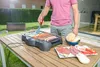 A person is grilling food on an electric grill on a wooden table outdoors. Náhled obrázku produktu