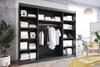 A bedroom scene featuring a large black wardrobe with sliding doors, filled with neatly folded clothes and storage baskets. A plant and a wicker chair with a yellow cushion are visible in the background. Produktbild-Vorschau