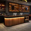 A modern bar counter with wooden panels and integrated LED lighting, set against a dark gray wall with shelves of bottles behind it. Produktbild-Vorschau