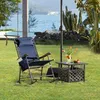 A blue rocking chair and a small table with flowers and fruit are set up on a grassy lawn by the water, with a swing set in the background. Produktbild-Vorschau