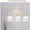 A three-light pendant lamp with white shades and wood accents hangs against a textured concrete wall. Náhľad obrázku produktu