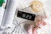 A digital alarm clock is placed on a bed next to a magazine and flowers. Podgląd zdjęcia produktu