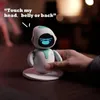 A hand holding a small, white and teal robot with a blue screen face, sitting on a charging base on a wooden table. Náhled obrázku produktu
