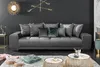 A large, plush gray sofa with multiple decorative pillows is arranged in a modern living room setting, with coffee tables and a rug in the foreground. Produktbild-Vorschau