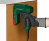 A gloved hand is holding a green electric tacker and using it on a brown particle board panel, which is attached to a white cabinet. Produktbild-Vorschau