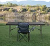 A folding table and chair set is set up on a grassy area next to a body of water, with hills and mountains in the background. Produktbild-Vorschau