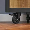 Close-up of a black caster wheel with a brake lever attached to the bottom of a furniture item with a wood-grain finish. Produktbild-Vorschau