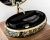 A black oval countertop sink with gold ornate detailing and a gold faucet, placed on a wooden surface. Produktbild-Vorschau