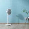 A white standing fan is placed on a wooden floor in front of a light blue wall. A small plant in a vase sits on a round side table to the right. Anteprima dell’immagine prodotto