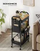 A black rolling cart with a wood-tone top shelf is filled with various items, including a coffee carafe, a plant, a phone, books, and storage containers. It is placed on a rug next to a sofa. Produktbild-Vorschau
