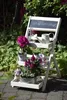 A white, foldable plant stand with three tiers, decorated with flowers and gardening accessories, set outdoors in a garden. Produktbild-Vorschau