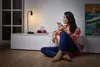 A woman is sitting on the floor in a living room, holding a smartphone and looking away from the camera. A lamp and decorative items are on a cabinet behind her. Produktbild-Vorschau