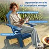 A woman sits in a blue Adirondack chair on a patio, reading a magazine. A fire pit is visible to the right. Produktbild-Vorschau