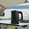 A black electric kettle is placed on a table outdoors, with a person in the background and a body of water behind them. Produktbild-Vorschau