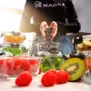 A person is shown preparing food by placing blueberries into a glass container, surrounded by other containers filled with fruits and vegetables. Produktbild-Vorschau
