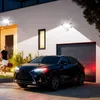 A man walks towards a car parked in front of a modern house with outdoor lighting. Produktbild-Vorschau
